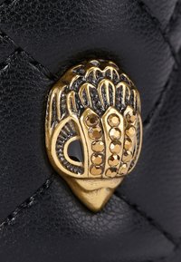 Czarna skórzana powierzchnia z złotym dekoracyjnym zapięciem, które ma skomplikowane detale i małe, zaokrąglone ćwiekami. Teksturowany, wypukły wzór akcentuje całość.