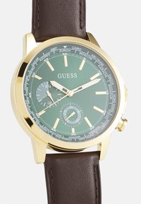 Orologio con finitura oro, quadrante verde, cinturino in pelle marrone, sub-quadrante dei secondi e nomi delle città contrassegnati sulla lunetta per le impostazioni del fuso orario mondiale.