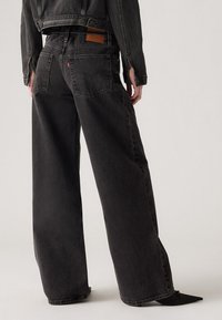 Svarte vide jeans med høy midje, laget av denim. Har en lærpakke på baksiden av midjebåndet og to baklommer.