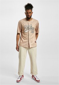 Jersey de baseball bej cu dungi albe, inscripționat cu litere negre "FUBU 05," cu nasturi în față, purtat cu pantaloni de culoare deschisă și adidași roșii.