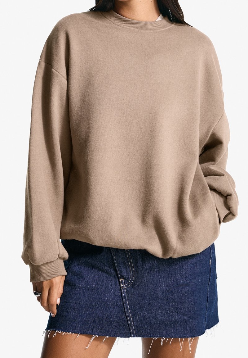 Kobieta ubrana w beżowy, oversize'owy sweter z prążkowanymi mankietami i obrębem, w połączeniu z postrzępioną, ciemnoniebieską mini spódniczką dżinsową.