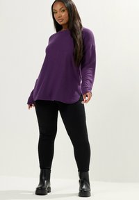 Top viola a maniche lunghe con orlo arrotondato, abbinato a leggings neri e stivaletti neri alla caviglia. Il tessuto sembra morbido e leggermente texturizzato.