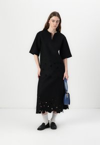 Svart klänning med korta ärmar, med utskurna blommönster vid fållen. Modellen håller en blå handväska, i kombination med vita strumpor och svarta skor.