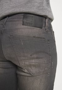 Jeans de mezclilla gris con un acabado texturizado, que cuentan con un parche de cintura de piel negra y detalles sutiles desgastados en los bolsillos.