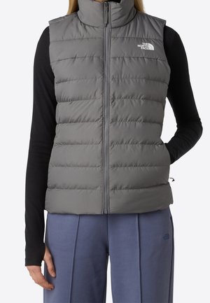 Femme portant un gilet matelassé gris avec une fermeture éclair à l'avant, un haut à manches longues noir et un pantalon bleu, les mains détendues le long du corps.