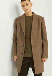 Abrigo de lana marrón con solapas en pico, tres botones y corte recto, superpuesto a un suéter de punto texturizado verde oscuro y pantalones de pana marrones.