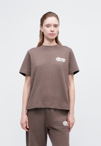 Sieviete, kas valkā brūnu brīva piegriezuma T-kreklu un bikses ar baltu "ellesse" logotipu uz krūšu un augšstilba, pret vienkāršu gaišu fona.