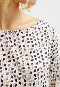 Top bianco con motivo geometrico blu navy e beige. Tessuto morbido, scollatura a barca e maniche corte. Focus sulle forme uniche nel design.