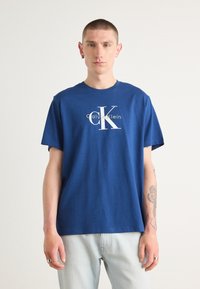 Camiseta de algodón azul con un gran logo blanco "CK" y el texto "Calvin Klein", cuello redondo y mangas cortas. Combinada con jeans claros.