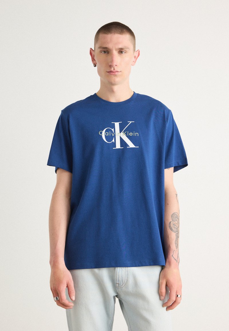 Camiseta de algodón azul con un gran logo blanco "CK" y el texto "Calvin Klein", cuello redondo y mangas cortas. Combinada con jeans claros.