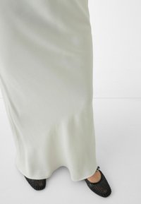 Lýtko v dlouhé hladké béžové sukni a černých tkaných balerínách na jednoduchém bílém podlaze a pozadí stěny.