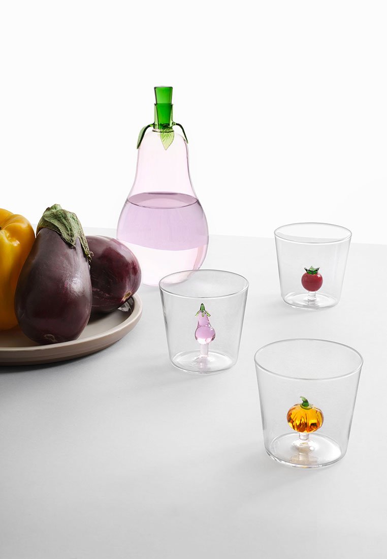 Przezroczyste szklanki do picia z podstawami w kształcie owoców: różowa bakłażan, czerwona pomidor i pomarańczowa dynia. Obecny jest również zielony szklany karafka i warzywa.