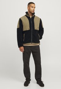 Giacca in fleece con pannelli neri e verde oliva a contrasto, chiusura con zip, colletto alto e tasche laterali. Indossata con pantaloni e scarpe scure.