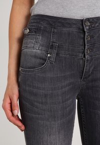 Šedé džíny z denimu s vysokým pasem, se spoustou knoflíkových zapínání, předními kapsami a přiléhavým střihem. Hladká textura s jemným vyblednutím.