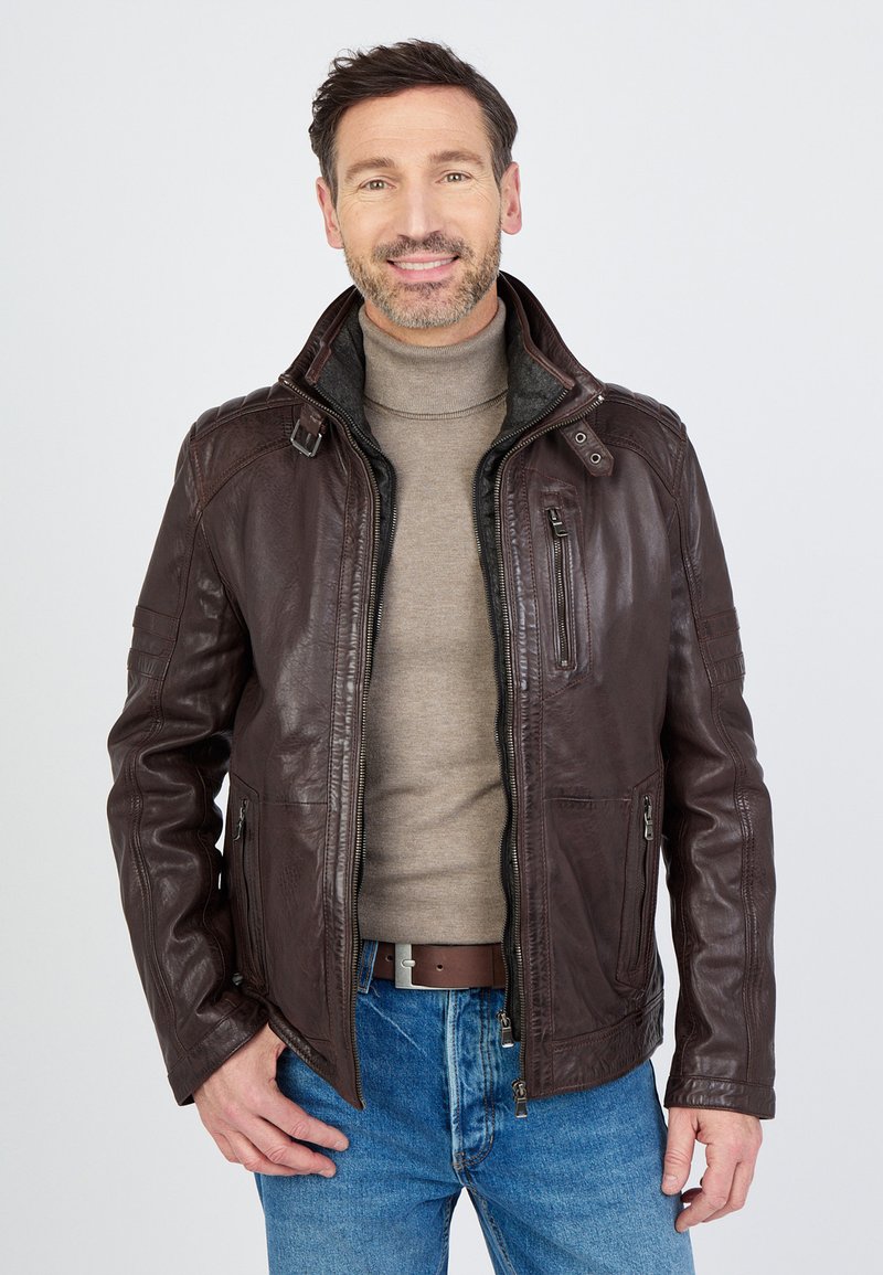 Bruine leren jas met ritszakken, hoge kraag en gestructureerde naden, gedragen over een grijze coltrui, in combinatie met blauwe jeans.