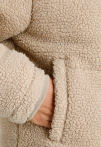 Grietos spalvos pūkų striukė su minkštu, tekstūruotu paviršiumi. Turinti paslėptą kišenę ir įpjautas rankoves, užtikrinančias tvirtą prigludimą.