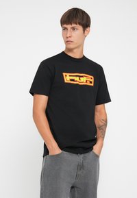 Black cotton T-shirt featuring a bold red and yellow logo design on the front. Short sleeves and a crew neckline. Model wears gray denim jeans.