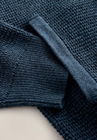 Sötétkék kötött pulóver, texturált szövettel és bordás mandzsettákkal, valamint derékrésszel. Az anyag lágy, sűrű és strukturált mintázatú.