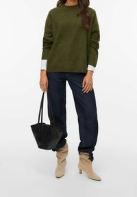 Personne portant un pull vert olive, un jean bleu foncé, des bottines beiges décontractées, tenant un sac fourre-tout en cuir noir sur le côté.