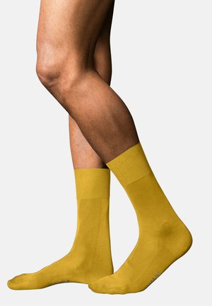 Piernas cruzadas de una persona con calcetines amarillo brillante hasta la mitad de la pantorrilla, de pie contra un fondo claro y liso.