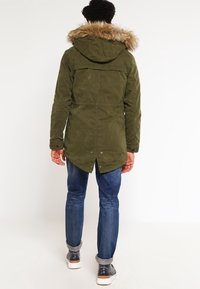 Olivgrön parka-jacka med en foder av päls på huvan, justerbart dragsko i midjan och en längre bakre fåll, i kombination med blåjeans och sneakers.