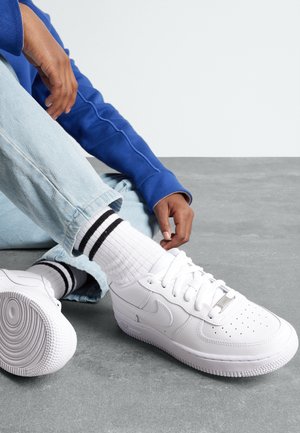 Zapatillas blancas Nike Air Force 1 con puntera perforada, cordones planos y suela de caucho texturizada. Se llevan con calcetines blancos con rayas negras.