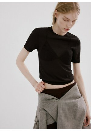 Persona joven ajustando una americana gris atada a la cintura, con una camiseta negra de manga corta con patrones triangulares texturizados en la parte delantera.