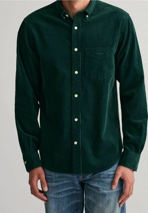 Homme portant une chemise en velours côtelé vert foncé à boutons avec une poche poitrine et un jean bleu, debout avec les mains détendues le long du corps.