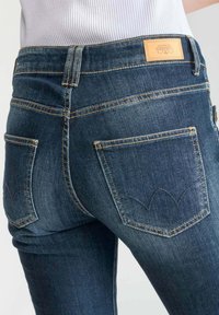 Jeans en denim bleu foncé avec une coupe slim, dotés de surpiqûres doubles et de deux poches arrière avec un motif de couture. Patch de marque en cuir sur la ceinture.