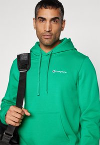 Felpa verde con cappuccio in cotone, dotata di cappuccio regolabile con cordino, tasca frontale e piccolo logo ricamato bianco e rosso sul petto.