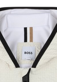 Capuche en tissu texturé blanc avec bord noir, ornée d'un accent rayé noir et blanc et d'une étiquette logo visible portant la mention "BOSS."