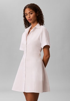 Kvinde med krøllet hår iført en kortærmet, hvid kjole med krave og knapper foran, stående mod en ensfarvet grå baggrund.