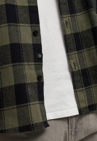 Camisa de franela a cuadros verdes y negros, con diseño de botones. Debajo, hay una camiseta blanca sencilla, con pantalones grises visibles.