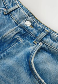 Primer plano de un pantalón vaquero azul mostrando el botón frontal, los trabillas para el cinturón y las costuras detalladas en la cintura y el área del bolsillo.