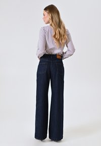 Wijd gesneden denim jeans in donkerblauw met een gestructureerde afwerking. Voorzien van achterzakken en een leren merkpatch op de taille. Verticaal gestreefd shirt gedragen.