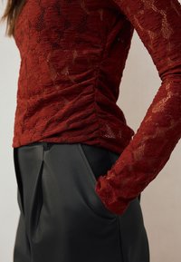 Top rosso in pizzo a maniche lunghe con dettaglio arricciato sul lato, abbinato a pantaloni neri in ecopelle a vita alta con pieghe marcate e tasche.