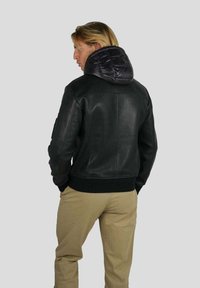 Homme aux cheveux blonds portant une veste en cuir noire avec une capuche matelassée et un pantalon beige, se tenant contre un fond clair.