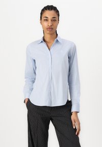 Camicia a righe blu e bianche con maniche lunghe, tessuto morbido e orlo curvo, indossata con pantaloni neri a righe sottili. Nessun logo visibile.