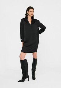 Robe noire avec un col en V et des manches longues, fabriquée dans un tissu lisse. Portée avec des bottes noires montantes jusqu'aux genoux. Design simple et épuré.