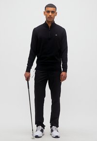 Άνδρας με μαύρο πουλόβερ με φερμουάρ στο λαιμό και παντελόνι, κρατώντας ένα μπαστούνι του γκολφ, στέκεται μπροστά σε ένα απλό ανοιχτό φόντο.