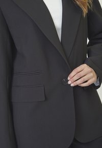 Tailored black blazer with a smooth texture, broad lapels, two front pockets, and a black button for closure. White undershirt visible.