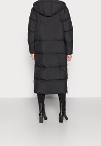 Lång svart puffjacka med huva, med quiltade partier, rak silhuett och dragkedjedetaljer vid ärmsluten, kombinerad med svarta stövlar.