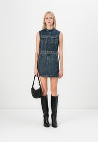 Denim shirt dress in dark blue with sleeveless design, button front, pockets, and belted waist. Black knee-high boots and studded handbag.