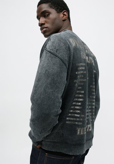 Sweatshirt gris foncé avec une texture délavée, affichant des noms de villes imprimés dans le dos. Poignets et ourlet côtelés. Coupe décontractée.
