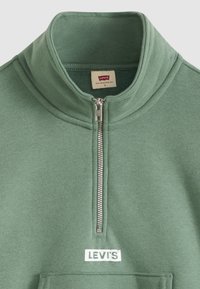 Polaire verte à col demi-fermeture éclair avec une poche poitrine. Dispose d’une fermeture éclair argentée et d’un écusson blanc avec le logo Levi’s.