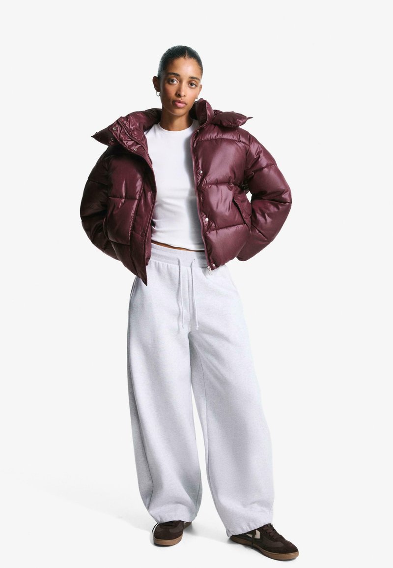 Bordeaux pufferjack met een hoge kraag, gestructureerd oppervlak; gecombineerd met lichtgrijze sweatpants. Bruine sneakers maken de outfit compleet.