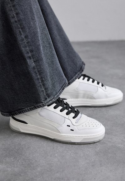 Witte sneakers met zwarte veters en mesh-details. Beschikt over een gladde leren bovenkant en een gestructureerde rubberen zool. Gepaard met brede grijze jeans.