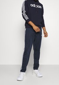 Hoodie Adidas bleu marine avec logo blanc, associé à un pantalon de survêtement bleu marine avec des rayures blanches. Les chaussures sont blanches avec un design à lacets.