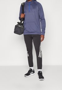 Felpa blu con tasca frontale e logo stampato. Pantaloni neri con strisce bianche sui lati. Scarpe nere e borsa a tracolla grigia.