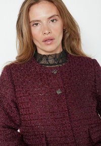 Burgundi tekstuuriga jakk, millel on kangast muster, kahte metallic-nuppu ja must pitsist kaelus all.